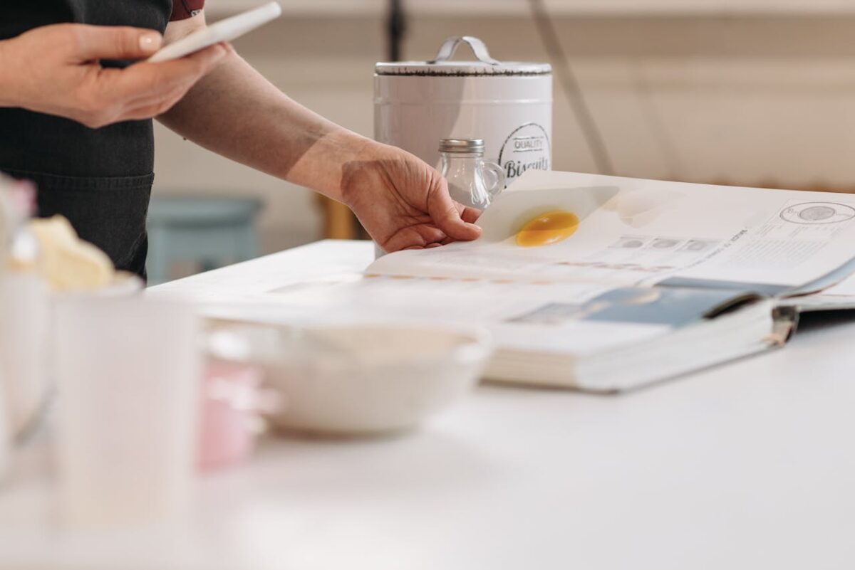 Person reading a recipe book with baking essentials in a kitchen setup.
